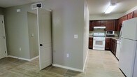 This single-story space presents a straightforward and functional layout, as seen through the open sightline connecting the entryway corridor to the kitchen. The flooring throughout consists of large-format light-toned tiles, lending a clean and cohesive look to the space. The walls are finished in a neutral greige tone, complemented by crisp white baseboards and door trim that provide a subtle contrast.

A paneled interior door with a dark handle is visible along the corridor, indicating separation between rooms or utility areas. The kitchen, positioned at the end of the hallway, features rich dark wood cabinetry that extends to the upper walls, providing ample storage. A white freestanding range sits beneath a ventilation hood, and the countertops appear to feature a subtle pattern, likely a laminate or stone-look surface. Overhead cabinetry frames the cooking area efficiently.

Ceiling-mounted HVAC vents are present throughout, suggesting a central air system. Electrical outlets are placed at standard heights along the walls. The overall ambiance is practical and move-in ready, offering a neutral palette well-suited for a variety of interior styles.