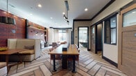 This thoughtfully designed business center spans a single floor and offers a variety of functional zones tailored for productivity and collaboration. The main open area anchors the space with a substantial communal table surrounded by leather-accented chairs, illuminated by a striking geometric pendant light fixture overhead. The herringbone-patterned carpet runs throughout, lending a sophisticated yet warm aesthetic to the entire floor.

A dedicated workstation alcove features built-in desks equipped with computers and a printer, set against a dramatic textured brick-red accent wall adorned with Edison-style pendant lights and open shelving — creating an inspiring, industrial-chic backdrop for focused work.

A private conference room, accessible through a handsome sliding barn-style wood door with glass panels, offers a more intimate setting. Inside, a butcher-block wooden table is surrounded by upholstered chairs, complemented by a wall-mounted flat-screen display, framed nature-inspired artwork, and a gold pendant light. Built-in cable ports in the table support seamless connectivity.

The corridors and transition areas are curated with framed artistic prints and sculptural wall pieces, reinforcing the space's design-forward character. Natural light filters in through multiple windows dressed with patterned curtains, while recessed lighting ensures consistent illumination throughout.