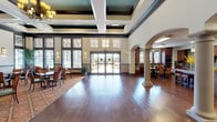 This community room and kitchen is a well-appointed shared space designed to accommodate a variety of social gatherings and events. The main hall is immediately striking, with soaring coffered ceilings finished in teal and dark wood trim, an elegant chandelier, and abundant natural light streaming through large clerestory windows and full-length glass doors that open to an outdoor patio area. Rich hardwood flooring extends throughout, transitioning to patterned area rugs in the dining seating zone, where round tables and upholstered chairs are arranged for comfortable group dining. Decorative arched columns add architectural character and help define the open floor plan. The fully equipped kitchen is a standout feature, outfitted with warm-toned wood cabinetry, granite countertops, a large central island with ornately turned legs, stainless steel appliances including a refrigerator, dishwasher, microwave, and wall oven, and a decorative tiled backsplash. A bank of arched windows above the kitchen sink floods the workspace with natural light. Pendant lighting and open display shelving add both function and style. The space connects seamlessly between the main gathering hall and kitchen via an arched passageway, creating a cohesive and inviting environment suited for community events, celebrations, and daily social interaction.