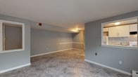This single-floor unit offers a practical and cohesive layout finished in cool grey tones with crisp white trim throughout. The main living area is generously sized, featuring neutral marble-patterned tile flooring, a pass-through window connecting to the kitchen, and baseboard heating. The open design allows for flexible furniture arrangements, making the space feel airy and versatile.

The kitchen is well-equipped with white shaker-style cabinetry, granite-look countertops, a stainless steel sink, an electric range, and a refrigerator. A decorative mosaic tile backsplash adds a touch of visual interest and ties the space together neatly.

Two bedrooms are carpeted in neutral tones, providing warmth underfoot. Each room is painted in the same soft grey palette and includes baseboard heating. One bedroom features a closet, offering practical storage.

The bathroom is finished with white tile walls and matching tile flooring set in a diagonal pattern. It includes a toilet, vanity, and towel bar, presenting a clean and functional space.

Throughout the unit, consistent finishes — including the tile flooring, grey paint, and white trim — create a unified and move-in ready aesthetic. The floor plan flows logically from the living area to the private rooms, balancing openness with defined spaces.