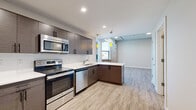 This 2-bedroom, 1-bathroom unit blends industrial character with modern finishes across a single, well-organized floor. The open kitchen serves as a central feature, equipped with stainless steel appliances including a range, microwave, and dishwasher, complemented by dark wood-textured cabinetry, white quartz countertops, and a white subway tile backsplash. Pendant lights with yellow accents add a contemporary touch, while wood-look plank flooring flows consistently throughout the unit.

The living area is a standout space, showcasing exposed brick walls, white-painted structural beams on the ceiling, and multiple large windows that allow ample natural light and outward views. The combination of raw brick and refined finishes creates a distinctive loft-inspired atmosphere.

The spacious primary bedroom carries the same wood-look flooring and features exposed ductwork along the ceiling, reinforcing the industrial aesthetic. The second bedroom offers a clean, neutral design with the same flooring. The bathroom is fitted with a sliding glass-enclosed walk-in shower with white tile surround. A hallway with built-in shelving provides additional storage throughout the unit.