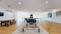 Located at 15 Abbey Lane in Danbury, CT, this single-floor clubhouse is designed with an open, airy layout that prioritizes community gathering and versatile use. The expansive main lounge serves as the heart of the space, featuring light hardwood flooring, recessed lighting, and white walls adorned with colorful framed artwork. A central seating arrangement — including a bold blue velvet sofa, accent chairs, and a wood coffee table — anchors the room, while high-top communal tables provide flexible gathering areas. A fireplace with a wall-mounted television above it anchors one end of the lounge, flanked by generous windows that frame lush tree-lined views and flood the interior with natural light. French doors along the perimeter provide seamless access to the outdoors. A dedicated conference room off the main area features a large rustic wood table, modern green chairs, and a wall-mounted display screen, making it well-suited for meetings or private events. The welcoming entry foyer offers built-in cabinetry, additional seating, and glass-paned double doors that invite natural light from multiple directions. Throughout the space, crown molding and clean architectural lines contribute to an overall atmosphere that is both polished and approachable.