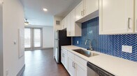 This single-floor unit offers a thoughtfully designed layout with a clean, contemporary aesthetic carried throughout each space. The kitchen serves as a visual centerpiece, showcasing crisp white shaker cabinetry, light quartz countertops, and a striking navy blue herringbone tile backsplash that adds bold character to the room. Stainless steel appliances, including a refrigerator, range, microwave, and dishwasher, provide a sleek, functional finish. Rich wood-style flooring runs continuously throughout the unit, grounding the space with warmth and visual cohesion.

The open living area adjoins the kitchen seamlessly, creating a spacious, flexible environment well-suited for both relaxing and entertaining. Natural light filters in through a large sliding glass door, offering views to the exterior and brightening the living space. The bedroom is generously proportioned, featuring both a wide window and a full glass door that allow ample natural light to fill the room.

The bathroom features a gray-toned vanity with a white quartz countertop and a large framed mirror, complementing the overall modern palette. Directly accessible from the bathroom, a walk-in closet provides notable storage capacity with built-in shelving and hanging rods, adding a practical and desirable element to this well-appointed unit.