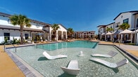 Located at 1230 Front Pl in North Port, Florida, this resort-inspired outdoor amenity space delivers a compelling blend of Mediterranean-influenced architecture and modern recreational design. The centerpiece is a generously sized swimming pool with crystal-clear turquoise water, complete with a water slide, wide sun shelves, and a surrounding deck outfitted with lounge chairs and umbrella-shaded seating. Adjacent to the main pool, a dedicated spa and hot tub offers a more intimate relaxation experience.

Multiple white-canopied cabanas line the pool deck, each furnished with plush round daybeds and draped curtains, providing shaded retreats for private lounging. Overhead string lights weave throughout the outdoor area, lending a warm ambiance during evening hours. A covered outdoor grilling station with a pergola-style roof provides a functional and social cooking space set against manicured green lawns.

The surrounding multi-story buildings feature white stucco facades, dark-trimmed balconies, and terracotta-accented rooflines consistent with a coastal Mediterranean aesthetic. Tall palm trees are thoughtfully planted throughout the grounds, reinforcing the Florida resort atmosphere. The space is enclosed by sleek black iron fencing, ensuring a sense of privacy while maintaining open, scenic sightlines across the well-kept landscape.