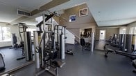 This fitness center spans two floors and is designed to support a comprehensive workout experience. The main floor is outfitted with a variety of strength-training equipment, including a multi-station cable machine and several resistance machines, providing users with options for full-body workouts. The durable, dark rubberized flooring offers both safety and comfort underfoot throughout the exercise space.

Large mirrors line the walls, allowing users to monitor their form while training and visually expanding the sense of space within the facility. The vaulted ceiling rises prominently above the main floor, enhancing the open, airy atmosphere of the room. Recessed lighting provides consistent, even illumination throughout.

Motivational fitness-themed artwork is displayed on the upper wall area, contributing to an energizing ambiance. A staircase leads to the second floor, which appears to offer additional equipment and training areas, making the facility well-suited for varied fitness routines. Natural light filters in through windows near the entry, brightening the space and creating a welcoming environment. Overall, this fitness center is thoughtfully arranged to maximize functionality while maintaining a clean, organized aesthetic.