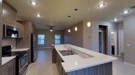 This single-floor space showcases an open-concept layout that seamlessly connects the kitchen and living areas, creating a sense of spaciousness and ease of movement throughout. The kitchen is a standout feature, anchored by an expansive island with a light-toned quartz countertop that offers generous workspace and casual seating. A double stainless steel sink is integrated into the island, accompanied by modern fixtures. The cabinetry features a warm wood-toned finish, complementing the light-toned luxury vinyl plank flooring that extends throughout the space. Three elegant pendant lights hang above the island, providing focused task lighting while adding a contemporary design element. Recessed lighting is distributed across the ceiling for consistent ambient illumination. The adjoining living area benefits from natural light streaming through multiple windows and is equipped with a ceiling fan for added comfort. A stainless steel refrigerator anchors one end of the kitchen. Neutral wall tones throughout contribute to a clean, modern aesthetic, while a doorway visible in the background hints at additional spaces beyond the main living area.