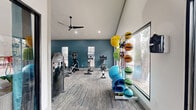 This fitness center offers a comprehensive workout environment designed to accommodate a wide range of exercise routines. The space is defined by its striking vaulted ceilings, which lend an airy, open feel throughout, while multiple ceiling fans ensure comfortable air circulation during workouts. Patterned carpet tile flooring runs the length of the facility, and bold teal-blue accent walls provide a modern, energizing aesthetic against the crisp white ceiling.

The cardio zone is well-appointed with a lineup of treadmills, elliptical trainers, and stationary bikes, several equipped with individual display screens. Adjacent to the cardio area, a dedicated strength training section houses a multi-station cable machine, a weight bench, and a barbell rack with plate weights. A full-length wall mirror spans a significant portion of the space, enhancing the sense of openness and allowing users to monitor their form.

A functional training area rounds out the facility, featuring a rack of medicine balls, foam rollers, and resistance equipment for flexibility and conditioning work. Large windows along the perimeter invite ample natural light and offer views of the surrounding outdoor environment. Exposed ductwork along the upper walls adds an industrial design touch, complementing the overall contemporary style of this thoughtfully outfitted fitness center.
