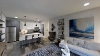 This single-floor residence showcases a thoughtfully designed open-concept layout that seamlessly connects the kitchen and living areas into one cohesive, modern environment. The kitchen is outfitted with crisp white cabinetry, dark granite countertops, and stainless steel appliances, anchored by a breakfast bar with white bar stools and illuminated by a trio of sleek pendant lights. The polished concrete floors extend throughout the space, lending an industrial-chic character that ties the design together. The living area is warm and inviting, centered around a neutral sofa and a striking patterned area rug in navy and white. A built-in shelving niche along the wall provides both functional storage and a curated display space for décor and books. A bold large-scale blue and gray artwork commands attention on the adjacent wall, adding a dramatic focal point. A sculptural dark accent chair adds contrast and visual interest to the sitting area. The overall ambiance is contemporary and refined, with a cool, neutral palette of grays and whites accented by deep navy tones, creating a sophisticated and cohesive aesthetic throughout the space.