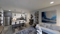 This single-floor residence showcases a thoughtfully designed open-concept layout that seamlessly connects the kitchen and living areas into one cohesive, modern environment. The kitchen is outfitted with crisp white cabinetry, dark granite countertops, and stainless steel appliances, anchored by a breakfast bar with white bar stools and illuminated by a trio of sleek pendant lights. The polished concrete floors extend throughout the space, lending an industrial-chic character that ties the design together. The living area is warm and inviting, centered around a neutral sofa and a striking patterned area rug in navy and white. A built-in shelving niche along the wall provides both functional storage and a curated display space for décor and books. A bold large-scale blue and gray artwork commands attention on the adjacent wall, adding a dramatic focal point. A sculptural dark accent chair adds contrast and visual interest to the sitting area. The overall ambiance is contemporary and refined, with a cool, neutral palette of grays and whites accented by deep navy tones, creating a sophisticated and cohesive aesthetic throughout the space.