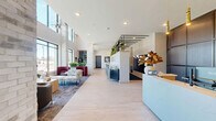 Located at 705 1st Ave SW in Rochester, Minnesota, this contemporary lobby presents a thoughtfully designed first-floor space that combines functionality with refined aesthetics. The lobby is defined by soaring double-height ceilings, light-toned hardwood flooring, and stacked stone brick columns that add architectural character throughout.

At the entrance, a sleek white reception desk anchored by gold pendant lighting and a rich, dark wood accent wall creates a polished focal point. Adjacent to the reception area, a communal bar-style counter with wire metal barstools sits beneath an open black metal shelving unit adorned with lush potted plants and framed artwork, blending greenery with urban design sensibilities.

Two distinct lounge areas offer comfortable seating arrangements. One features bold burgundy rounded chairs paired with a low circular coffee table, set atop a patterned area rug beneath floor-to-ceiling windows that flood the space with natural light and provide open views of the surrounding streetscape. A second lounge area offers soft blue upholstered chairs and a natural wood console, creating a calmer, more intimate setting. A large-scale floral mural adds a vibrant artistic statement to the interior. The overall ambiance is modern, welcoming, and carefully curated.