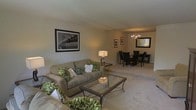 This single-floor space offers a seamless, open-concept layout that connects the living and dining areas into one cohesive, inviting environment. The living area is anchored by a plush, neutral-toned sofa adorned with decorative green and white accent pillows, complemented by a glass-top coffee table with ornate metal legs that adds a refined touch. A matching side table with a classic table lamp provides warm, intimate lighting, while a framed black-and-white bridge photograph serves as a striking focal point on the wall. The space flows naturally into the dining area, where a round table is surrounded by dark wood chairs, positioned beneath an elegant chandelier that casts a soft, welcoming glow. A large rectangular mirror on the dining area wall enhances the sense of depth and brightness. The flooring throughout is covered in wall-to-wall neutral carpeting, contributing to a cohesive and comfortable atmosphere. Soft, warm-toned walls tie the entire space together, creating an ambiance that is both functional and stylish. The overall design reflects a classic, tastefully curated aesthetic suitable for everyday living and entertaining.