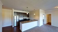 This single-floor space showcases an open-concept layout that integrates the kitchen and living areas into a cohesive, functional environment. The kitchen is a standout feature, equipped with stainless steel appliances including a full-size refrigerator, microwave, and range. Rich, dark cabinetry provides ample storage and contrasts elegantly with the granite-topped peninsula, which includes an integrated sink and offers additional seating or prep space. Pendant lighting hangs above the peninsula, adding focused illumination and a contemporary design touch, while a track lighting system above the main cabinetry ensures the workspace is well-lit. The kitchen transitions naturally into the carpeted living area, creating a sense of openness and easy movement throughout the space. Neutral wall tones complement the dark wood-finish cabinetry and granite surfaces, contributing to a clean, modern aesthetic. A hallway visible in the background leads to additional rooms, with a windowed door hinting at natural light filtering through from adjoining spaces. The overall ambiance is modern and practical, balancing style with everyday functionality.