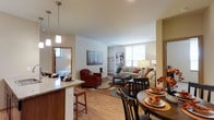 This single-floor residence is situated within a contemporary multi-story building that combines stone, wood siding, and modern metal accents for a polished exterior aesthetic. The building features expansive outdoor patio spaces furnished with seating areas, large planters with seasonal flowers, and railed walkways overlooking lush green surroundings.

Inside, the unit showcases an open-concept layout where the kitchen, dining area, and living room flow seamlessly together. The kitchen features a granite-topped breakfast bar with bar stool seating and warm wood cabinetry, with pendant lighting overhead adding a functional yet stylish touch. Light hardwood flooring extends throughout the main living areas, contributing to a bright, cohesive feel.

The dining area accommodates a full-sized table and chairs, positioned conveniently adjacent to the kitchen. The living room is generously proportioned, centered around a coffee table and area rug, with a built-in shelving unit along one wall and a large window allowing ample natural light. A glimpse into the bedroom reveals a comfortable, well-lit space. Overall, the unit presents a warm, well-designed environment with a welcoming ambiance.