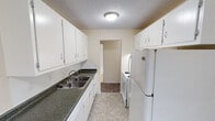 This single-floor residential unit offers a practical and well-organized layout designed for comfortable everyday living. The galley-style kitchen is outfitted with white cabinetry providing ample storage, dark granite-look countertops, a double stainless steel sink, and white appliances including a refrigerator and range. Light tile flooring runs throughout the kitchen, which opens into a dedicated dining area with additional cabinet storage and a window that brings in natural light. The living room is generously sized, featuring neutral-toned carpet, textured ceilings, and baseboard heating. A large window with vertical blinds allows natural light to fill the space, and a wall-mounted air conditioning unit provides additional climate control. The entry door opens directly into this open living area, creating an inviting flow. Two bedrooms continue the carpeted flooring and warm neutral wall tones, each with windows overlooking the surroundings and baseboard heating for year-round comfort. The full bathroom features tile flooring, a combined tub and shower with white subway tile surround, a toilet, and a vanity with under-sink storage and an overhead medicine cabinet. The overall aesthetic is clean and straightforward, offering a move-in-ready space with consistent finishes throughout.
