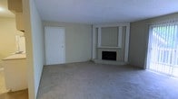 This single-floor residential space offers a straightforward and functional layout designed for comfortable everyday living. The living area serves as the heart of the home, immediately drawing attention with its built-in fireplace framed by a paneled accent wall and a white mantel surround. The fireplace is set into a raised hearth, adding a focal point that brings warmth and character to the room. Neutral, light-toned walls complement the soft, plush carpeting that extends throughout the space, creating a cohesive and understated aesthetic. The entry door, finished in crisp white with polished gold hardware, provides a clean and welcoming transition into the living area. The walls are painted in a muted, warm beige, contributing to the calm and inviting ambiance of the interior. The ceiling is smooth and white, helping to reflect light and maintain an open feel throughout the room. Overall, the space presents a quiet, neutral palette that offers a versatile canvas, well-suited for a range of furnishing styles and personal touches.