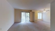 This single-floor space offers a welcoming and well-lit interior defined by clean lines and a functional layout. The main living area is spacious and open, finished with neutral wall-to-wall carpeting that adds warmth underfoot. A large sliding glass door fitted with vertical blinds serves as a focal point, flooding the room with natural light while providing direct access to an outdoor area surrounded by lush greenery. An additional window on the adjacent wall further enhances the brightness of the space, ensuring a consistently airy atmosphere throughout. The walls are painted in a soft, warm beige tone that complements the light-filled environment and contributes to the overall sense of openness. A ceiling fan is visible in the adjoining area, offering both comfort and ventilation. The layout flows naturally between zones, making the space versatile and easy to navigate. Wall-mounted HVAC units are discreetly positioned to maintain climate control without interrupting the clean aesthetic. The combination of generous natural light, neutral finishes, and outdoor connectivity gives this space a calm, livable character well-suited to a variety of uses.