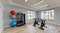 This fitness center is a comprehensively outfitted facility designed to support a full range of workout routines across multiple distinct zones. The space is organized into several interconnected areas, each tailored to a specific type of exercise.

The cardio and strength area features light-toned carpet flooring and is lined with large windows fitted with horizontal blinds, allowing ample natural light to pour in while offering views of palm trees outside. This zone houses treadmills, elliptical trainers, and stair climbers, with full-length mirrors along the walls that enhance the sense of space and allow users to monitor their form. A tiered dumbbell rack with a wide range of weights anchors the free weights section, accompanied by a flat bench.

A separate cycling studio provides a bright, dedicated space for spin workouts. It features light wood-look tile flooring, recessed tray ceilings with linear LED lighting, and multiple stationary cycling bikes arranged for group or individual sessions. A well-stocked equipment rack holds medicine balls in various weights, kettlebells, and stability balls, making it ideal for functional training.

The facility is finished with soft gray walls throughout, giving the entire space a clean, modern aesthetic. Thoughtful lighting, mirrored walls, and distinct flooring zones help define each workout area, creating a cohesive and functional environment for users of all fitness levels.
