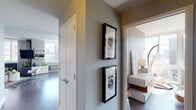 This thoughtfully designed single-floor residence offers a seamless blend of modern aesthetics and urban comfort. The open-concept living area is anchored by floor-to-ceiling windows that frame dramatic city skyline and waterfront views, flooding the space with natural light. Dark hardwood floors run throughout, complementing a neutral gray and white color palette accented with bold pops of color in the décor. The living room features a contemporary layout with a wall-mounted TV, built-in shelving, and a stylish mix of furnishings set atop a geometric area rug.

The kitchen is equipped with stainless steel appliances, warm wood cabinetry, a white quartz peninsula with bar seating, and pendant lighting — all framed by a window overlooking the water and city beyond. A dedicated sunroom-style nook with a hanging wicker chair offers a tranquil retreat with panoramic waterfront views.

Both bedrooms are well-appointed with large windows, generous natural light, and refined finishes. The primary bedroom features a tufted white upholstered headboard and curated artwork. The entry hallway is artfully decorated with framed black-and-white prints. Two bathrooms complete the layout — one with a glass-enclosed shower and wood-toned vanity, and another with a soaking tub and matching finishes.