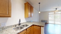 This single-floor unit offers a practical and cohesive layout centered around an open-concept kitchen and living area. The kitchen is well-appointed with warm honey-toned wood cabinetry, granite countertops, a double stainless steel sink, and black appliances including a range, over-the-range microwave, and dishwasher. A peninsula counter separates the kitchen from the living space, providing additional prep area and an open sightline throughout. Pendant lighting above the peninsula adds a functional decorative touch, complemented by track lighting near the entry.

Hardwood-style flooring runs consistently throughout the main living areas, lending warmth and visual continuity. The living area is spacious and bright, anchored by a ceiling fan and a large sliding glass door that invites natural light and provides outdoor access. Vertical blinds offer privacy and light control.

An additional room features the same flooring and neutral walls, offering flexible use as a bedroom or office. A dedicated closet with a built-in rack provides convenient storage near the entry.

The bathroom includes a wood-toned vanity with an integrated sink, a large mirror with vanity lighting, a toilet, and a shower-tub combination, all finished with the same warm hardwood-style flooring that unifies the overall aesthetic of the space.