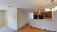 This single-floor residence offers a practical and comfortable layout designed for everyday living. The open-concept main area seamlessly connects the living and dining spaces, finished with warm hardwood-style flooring that transitions to plush carpeting in the lounge area. A chandelier near the entryway adds a touch of elegance, while the front door is flanked by an electrical panel and ample wall space. The kitchen is a standout feature, configured in a U-shape with warm honey-toned wood cabinetry, generous countertop workspace, a stainless steel sink, and a full suite of black appliances including a gas range, microwave, refrigerator, and dishwasher. A pass-through counter connects the kitchen to the main living area, enhancing the open flow of the space. The bedrooms are carpeted and well-sized, offering natural light through window-blinded windows. The primary bedroom is particularly spacious, benefiting from multiple windows that invite natural light. The bathroom is clean and functional, featuring tile flooring, a full bathtub and shower combo, and a bi-fold door that provides access to an adjacent laundry area. Throughout, neutral tones and white trim create a bright, cohesive aesthetic.