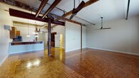 This single-floor studio showcases a compelling blend of industrial character and modern amenities. The expansive open living area is defined by richly patterned parquet wood flooring that extends throughout, while exposed ductwork and original iron pipes run overhead, lending an authentic loft aesthetic. Sturdy wooden support columns punctuate the space, further reinforcing its industrial heritage. Ceiling fans are strategically positioned across the open layout, complementing a wall-mounted mini-split system for efficient climate control. Natural light enters through windowed brick walls, highlighting the raw textural contrast between the masonry and crisp white painted surfaces.

The kitchen is well-appointed, featuring warm wood cabinetry, granite countertops, stainless steel appliances including a gas range, microwave, and refrigerator, and a peninsula with seating overhang illuminated by pendant and chandelier lighting. Adjacent to the kitchen, a built-in shelving and desk unit adds functional storage and workspace. A dedicated utility room houses a full-size washer and dryer alongside a water heater. The bathroom area features light-toned large-format tile flooring and wood vanity cabinetry with granite counters, providing a clean contrast to the industrial main living space.