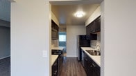 This single-floor residence offers a thoughtfully designed layout centered around a sleek galley-style kitchen. The kitchen is outfitted with rich, dark espresso cabinetry that extends from floor to ceiling on both sides, providing ample storage throughout. Light-toned countertops create a striking contrast against the dark cabinets, adding visual depth to the space. Stainless steel appliances — including a refrigerator, dishwasher, and built-in microwave — are complemented by a gas range cooktop, making the kitchen both functional and stylish. A stainless steel undermount sink with a modern faucet is conveniently positioned along the countertop. The kitchen is finished with warm wood-look flooring that runs continuously underfoot, adding warmth and cohesion. Overhead, a flush-mount ceiling light provides bright, even illumination. The textured ceiling adds a subtle architectural detail throughout. The kitchen opens with a clear sightline toward an adjacent room, allowing natural light to filter in and enhance the overall sense of openness. Neutral gray-toned walls throughout the space create a clean, versatile backdrop suitable for a variety of interior styles.