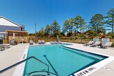 Located at 1551 Riverside Pkwy in Douglasville, Georgia, this outdoor amenity area is a well-appointed retreat designed for relaxation and recreation. The centerpiece of the space is a generously sized swimming pool with crystal-clear blue water, framed by a broad, light-toned concrete sundeck. The deck is furnished with a selection of cushioned lounge chairs and chaise lounges, providing ample space for sunbathing and socializing beneath the open Georgia sky. A separate, shallower wading area adjoins the main pool, adding versatility for residents of all ages. A stylish wooden pergola structure anchors one corner of the deck, offering a shaded gathering spot alongside outdoor seating and table arrangements. The pool area is enclosed by a black iron fence, lending both security and an open, airy feel that allows unobstructed views of the surrounding mature pine trees and green landscape. Adjacent to the pool, a covered porch area features board-and-batten siding in a bold navy and white color scheme, with additional seating for casual conversation or quiet relaxation in the shade. The multi-story residential buildings visible in the background echo the same contemporary coastal-inspired palette of navy blue and crisp white, creating a cohesive and attractive community aesthetic throughout the property.