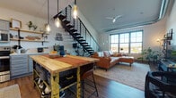 This two-floor urban loft showcases a striking industrial-modern aesthetic defined by soaring vaulted ceilings, exposed ductwork, and an open-concept layout that seamlessly connects the kitchen, dining, and living areas on the main level. A bold open-riser steel staircase with horizontal rail balusters serves as a dramatic architectural centerpiece, leading to the upper loft level. The main living area features rich hardwood flooring, large multi-pane windows that frame outdoor views and flood the space with natural light, and a ceiling fan overhead. Pendant jar lights hang above a butcher-block kitchen island with bar seating, complementing the well-appointed kitchen outfitted with gray shaker cabinetry, open wood shelving, white countertops, and stainless steel appliances. Two bathrooms echo the space's design language with gray vanities, white countertops, and wood-look flooring. The upper loft-level bedroom benefits from plush carpeting, skylights that bathe the room in daylight, a ceiling fan, and an open railing that offers a view down to the main floor. A second carpeted bedroom on the upper level also features vaulted ceilings and exposed ductwork, adding to the loft's cohesive industrial character throughout.