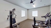 This fitness center is a dedicated single-floor exercise space designed to support a range of workout routines. The room is finished with neutral white walls accented by crown molding, lending a clean and polished look to the space. Durable carpet flooring covers the entire room, providing cushioning underfoot during workouts.

The equipment selection includes a multi-station strength training machine, a recumbent exercise bike, an elliptical trainer, and a treadmill, offering users options for both cardiovascular and resistance training. A large wall-mounted mirror spans a portion of one wall, visually expanding the space and allowing users to monitor their form during exercise.

Overhead, a ceiling fan with an integrated light fixture ensures adequate airflow and illumination throughout the room. Multiple windows fitted with horizontal blinds line the walls, inviting natural light while allowing occupants to control glare and privacy. A dedicated entry door marked with an exit sign provides clear access and egress, and posted fitness center rules are visible near the entrance, indicating this is a shared or community-use facility.

Overall, the space maintains a bright, functional ambiance suited for everyday fitness activities.