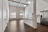 This 1-bedroom, 1-bathroom loft-style unit offers a compelling blend of historic industrial character and contemporary design. The open-plan living and kitchen area is anchored by original reclaimed timber columns and overhead wooden beam ceilings, which contrast beautifully against crisp white walls and modern hardwood flooring. Large arched windows along the exposed brick walls flood the living space with abundant natural light, casting dramatic shadow patterns throughout the day and enhancing the loft's airy, open atmosphere.

The kitchen is well-appointed with white shaker cabinetry, light subway tile backsplash, white quartz countertops, a center island with an undermount sink, stainless steel appliances, and stylish globe pendant lighting. Open shelving adds a functional yet modern touch.

The bedroom continues the industrial aesthetic with exposed brick, wooden ceiling planks, and generous natural light streaming through arched windows. The bathroom features classic white subway tile in the tub-shower surround, paired with marble-look floor tile and clean white vanity cabinetry. A dedicated closet provides practical storage. Throughout, the unit balances raw, historic materials with refined modern updates for a distinctive and inviting living environment.