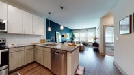 This thoughtfully designed single-floor residence showcases a seamless open-concept layout that connects the kitchen, dining, and living areas into one cohesive, light-filled space. The kitchen is fully equipped with stainless steel appliances, including a refrigerator, range, microwave, and dishwasher, complemented by granite countertops, two-tone cabinetry in white and warm gray, and a peninsula with bar seating illuminated by modern pendant lighting. Rich hardwood-style flooring extends throughout the main living areas, grounding the space with warmth and continuity. The living room is anchored by a bold deep-teal accent wall and oversized windows that flood the room with natural light and offer views of the outdoors. A dedicated dining area sits adjacent to the living space, enhancing the open, airy feel. The entry hallway features recessed lighting and clean white walls, providing a welcoming transition into the home. The bedroom offers plush carpeting, a striking burnt-orange accent wall, large windows with ample natural light, and a calm, restful atmosphere. The bathroom presents an elongated vanity with granite countertops, a walk-in glass-enclosed shower with subway tile, and hardwood-style flooring, delivering a spa-like experience with functional elegance.