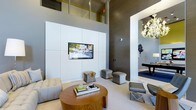 This single-floor space makes a striking impression through its dramatic double-height ceilings, which create an expansive, airy atmosphere that elevates the overall sense of scale. The main living area is anchored by a floor-to-ceiling white paneled media wall with a centrally mounted flat-screen television, flanked by tufted seating and a rich wood-toned round coffee table. Plush ottomans and accent chairs in neutral tones are thoughtfully arranged to encourage comfortable gathering. The walls feature a deep charcoal-toned finish that provides bold contrast against the crisp white architectural elements. An adjacent space, visible through a large open archway, reveals a darkly appointed room adorned with an ornate chandelier and a billiard or game table, adding an entertainment-focused dimension to the layout. Clean lines, high-end materials, and carefully curated décor define the contemporary design sensibility throughout. The interplay of light and shadow, enhanced by recessed and accent lighting, contributes to an atmosphere that is both refined and inviting, suited for both social gatherings and relaxed everyday use.