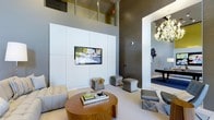 This single-floor space makes a striking impression through its dramatic double-height ceilings, which create an expansive, airy atmosphere that elevates the overall sense of scale. The main living area is anchored by a floor-to-ceiling white paneled media wall with a centrally mounted flat-screen television, flanked by tufted seating and a rich wood-toned round coffee table. Plush ottomans and accent chairs in neutral tones are thoughtfully arranged to encourage comfortable gathering. The walls feature a deep charcoal-toned finish that provides bold contrast against the crisp white architectural elements. An adjacent space, visible through a large open archway, reveals a darkly appointed room adorned with an ornate chandelier and a billiard or game table, adding an entertainment-focused dimension to the layout. Clean lines, high-end materials, and carefully curated décor define the contemporary design sensibility throughout. The interplay of light and shadow, enhanced by recessed and accent lighting, contributes to an atmosphere that is both refined and inviting, suited for both social gatherings and relaxed everyday use.