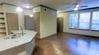 This single-floor space features an open-concept layout that integrates the kitchen and living areas into one cohesive environment. The flooring throughout is a warm-toned hardwood-style material that adds visual continuity and a polished look to the interior. The kitchen area is positioned to the left and includes a countertop with light granite or stone-like surfaces and a sink, providing a functional workspace that flows naturally into the main living area.

A central structural column or partial wall divides portions of the space, adding architectural definition without fully closing off the open floor plan. Two interior doors are visible, indicating access to additional rooms or storage areas. A window along the far wall allows natural light to filter in through slatted blinds, brightening the living area. A ceiling fan with a light fixture is mounted overhead, contributing to both comfort and illumination.

Crown molding along the upper walls and consistent neutral beige tones throughout give the space a clean, unified aesthetic. The overall layout is functional and straightforward, with a practical design that maximizes the use of the available floor area.