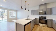 This thoughtfully designed 1-bedroom, 1-bathroom residence offers an efficient and modern layout across a single floor. The entrance welcomes you with light hardwood-style flooring and a glass-paneled door that allows natural light to filter in from an adjacent exterior corridor.

The kitchen is a standout feature, showcasing gray shaker-style cabinetry with matte black hardware, crisp white countertops, and a full suite of stainless steel appliances including a refrigerator with ice dispenser, electric range, built-in microwave, and dishwasher. A peninsula island with a single-basin sink and stylish pendant lighting above adds both function and visual appeal, creating a natural transition into the open living area.

The living room benefits from continued hardwood-style flooring and connects seamlessly to the kitchen, offering a cohesive open-concept feel. The bedroom is carpeted for comfort and features a window with blinds that invites natural light. A dedicated closet provides convenient storage.

The bathroom is finished with the same gray cabinetry as the kitchen, topped with a white vanity, a framed mirror, and warm overhead lighting, all set on hardwood-style flooring. A hallway connects the key areas of the home, completing a practical and well-organized floor plan.