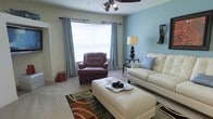 This single-floor space centers around a thoughtfully designed living room that combines comfort and style. The room is painted in a soothing light blue accent wall paired with neutral beige tones, creating a calm and welcoming atmosphere. Natural light flows in through a large window dressed with elegant grey curtain panels and white blinds, brightening the carpeted floor throughout. A ceiling fan with integrated lighting provides both air circulation and additional illumination. The seating area is well-appointed, featuring a cream-colored sofa accented with a blue throw pillow, a rich burgundy leather armchair, and a matching white rectangular ottoman that doubles as a coffee table. A patterned area rug in earthy tones of green, brown, and black anchors the seating arrangement. A sleek side table with a classic table lamp adds warmth to the corner of the room. Decorative wall accents and carefully placed accessories contribute to the polished, move-in-ready feel of the space. An open entryway visible on the left suggests a connected layout, enhancing the overall sense of flow within the home.