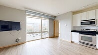 Located at 515 SE Huron Blvd in Minneapolis, MN, this one-bedroom, one-bathroom unit offers a smart and functional layout across a single floor. Light hardwood flooring runs throughout, lending warmth and continuity to the space. The open-concept main area seamlessly integrates the living room, dining space, and kitchen, creating a cohesive and efficient living environment. Large floor-to-ceiling windows flood the area with natural light and offer views of the surrounding Minneapolis neighborhood and cityscape beyond. An exposed duct runs along the ceiling, adding an industrial touch to the otherwise clean, modern aesthetic. The kitchen is equipped with white shaker-style cabinetry, dark countertops, subway tile backsplash, stainless steel appliances including a refrigerator, dishwasher, and built-in microwave, plus track lighting overhead. The bedroom is a well-proportioned retreat, also featuring a large window with outdoor views and access to a closet. The bathroom is finished with white walls, tile flooring, a dark-countered vanity with white cabinetry, a walk-in shower, and built-in shelving for added storage. This unit presents a clean, contemporary aesthetic well-suited for urban living in Minneapolis.