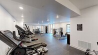 This fitness center offers a thoughtfully designed exercise space that accommodates a wide variety of workout routines. The room features clean white walls, recessed ceiling lighting, and durable rubber-tile flooring throughout, creating a practical and modern environment conducive to focused training.

The cardio zone is well-stocked with multiple Star Trac treadmills and elliptical machines, lined along one side of the room to maximize open floor space. A full-length mirror along one wall visually expands the space and supports strength and form-check exercises. Adjacent to the mirror, a rack of dumbbells and a weight bench provide ample free-weight training options.

Strength training is further supported by multiple cable and resistance machines positioned throughout the room, offering a broad range of muscle-targeting exercises. A dedicated area near the entrance features stationary bikes, a cross-trainer, and wall-mounted foam rollers, catering to flexibility and low-impact workouts. Stability balls are also available for core training.

Natural light filters in through several windows along the perimeter walls, offering views to the outside and keeping the space from feeling enclosed. The layout is open and efficiently organized, allowing multiple users to exercise simultaneously without congestion. Gym rules are posted on the wall, reflecting a well-maintained, community-oriented facility.