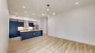 Located at 7716 Louis Pasteur Drive in San Antonio, TX, this thoughtfully designed single-story, 2-bedroom, 2-bathroom home offers a clean modern aesthetic with high-end finishes throughout. Light wood-tone flooring flows seamlessly across the open-concept living and kitchen area, creating a bright, cohesive feel. The living room is bathed in natural light from large windows and a glass door that opens to an outdoor view, and is accented by a stylish gold globe chandelier. The kitchen is a standout, featuring bold navy cabinetry, white quartz countertops, a marble-look backsplash, stainless steel appliances, a wall-mounted range hood, and a spacious island with seating. Both bathrooms feature glass-enclosed walk-in showers dressed in dramatic marble-look tile with herringbone-patterned floors, matte black hardware, and backlit vanity mirrors. The generously sized primary bedroom includes a ceiling fan, recessed lighting, and a sliding barn door leading to its closet. The second bedroom benefits from dual windows that bring in ample natural light. A hallway with clean-lined panel doors connects all rooms, complemented by matte black hardware details. The home also includes a dedicated laundry area and utility room for added convenience.