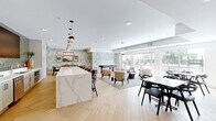 Located at 7689 Hardwood Ave S in Cottage Grove, Minnesota, this single-floor community space offers a thoughtfully designed open-plan layout that seamlessly connects a kitchen, dining room, and living area. Light hardwood-style flooring flows throughout, lending warmth and continuity to the entire space.

The kitchen is a standout feature, anchored by a large waterfall-edge quartz island with bar seating and pendant lighting overhead. White shaker cabinetry, sage green subway tile backsplash, stainless steel appliances, and a built-in wall-mounted display screen complete this well-appointed area.

The dining room is defined by nature-inspired wallpaper featuring a blue-toned pine tree motif that wraps the walls. A glass-top dining table seating eight sits beneath an industrial-style rectangular pendant light, creating an elegant yet inviting atmosphere.

The living area includes a wood-accented fireplace with a built-in TV surround and open shelving, flanked by a comfortable sectional and additional seating. A separate lounge zone offers plush armchairs arranged around a low coffee table, with oversized windows and glass doors providing abundant natural light and views of an outdoor patio. A billiards table adds a recreational element, making this a versatile and welcoming gathering destination.