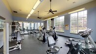 The Second Fitness Center is a spacious, single-floor exercise facility designed to support a comprehensive workout experience. The room is bathed in natural light, courtesy of large multi-pane windows lining multiple walls and offering views of the surrounding outdoor areas, including a pool and landscaped grounds. Ceiling fans suspended from the warm-toned ceiling provide airflow throughout the space, complementing the overhead fluorescent lighting that ensures consistent illumination.

The flooring consists of dark rubber matting, providing a safe and durable surface suited for a variety of exercises. A full-length mirror runs along one wall, visually expanding the space while allowing users to monitor their form during workouts.

The cardio section is well-stocked with treadmills, elliptical trainers, stationary bikes, and rowing machines, all positioned to take advantage of the natural light and outdoor views. The strength-training area includes a tiered dumbbell rack with a wide range of free weights, multiple weight benches, and cable machines for targeted muscle training. Additional selectorized strength machines are arranged along the walls, offering guided resistance options.

Overall, the fitness center presents a bright, functional, and well-organized environment suited for both cardio and strength training.