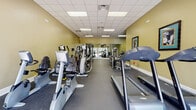 The fitness center at The Abbys At Inverness in Birmingham, Alabama, is a thoughtfully designed, single-level facility that provides residents with a comprehensive workout environment. The space features warm olive-toned walls complemented by a drop-panel ceiling with recessed fluorescent lighting, creating a bright and functional atmosphere throughout.

The facility is organized to accommodate a wide range of workout routines. A dedicated cardio zone lines one side of the main floor, housing multiple treadmills, elliptical trainers, and both upright and recumbent stationary bikes. Flat-panel televisions are mounted on the walls, allowing users to stay entertained during cardio sessions. Large windows at one end of the room invite natural light and offer outdoor views, enhancing the overall workout experience.

The strength training area is well-appointed with several cable and selectorized weight machines, catering to both upper and lower body workouts. A separate free weight alcove features dual racks of dumbbells in varying weights, paired with a flat workout bench and floor-to-ceiling mirrors for form monitoring. Additional mirrors are strategically placed throughout the space, amplifying the sense of openness. Durable rubber flooring covers the entire facility, providing both safety and comfort. A glass-panel entry and a climate control panel are also visible, reflecting attention to user comfort and accessibility.