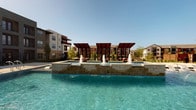 This contemporary amenity space is designed to deliver a resort-like experience, centered around a expansive outdoor pool with crystal-clear turquoise water. Three architectural pergolas with rich, dark wood slatted canopies rise above a raised stone-clad platform, each sheltering comfortable lounge seating. Decorative water fountains jet upward from the platform, adding a dynamic, elegant touch to the outdoor environment. The surrounding pool deck is finished in smooth concrete, offering ample space for lounging under the open sky.

The interior clubroom is equally impressive, boasting soaring double-height ceilings with warm wood paneling and floor-to-ceiling windows that frame unobstructed views of the pool area, flooding the space with natural light. A full-size pool table anchors the central gathering area, complemented by a shuffleboard table displayed alongside a trio of decorative woven textile wall hangings. Geometric diamond-pattern shelving units line the feature wall, adding a bold artistic element.

The kitchen area is a true standout, featuring a dramatic veined marble island, recessed wood coffered ceiling, pendant lighting, and a vibrant tiled accent wall combining teal, orange, and neutral tones. Dark cabinetry and professional coffee service complete the space, which opens seamlessly into the adjacent lounge through wide sliding glass doors.