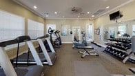 This fitness center offers a fully equipped, single-floor workout facility designed to support a wide range of exercise routines. The space features warm, neutral-toned walls accented by crisp white crown molding and baseboards, lending a polished, welcoming atmosphere. Recessed lighting throughout ensures the room is well-lit, complemented by natural light streaming in through multiple windows fitted with blinds. A ceiling fan promotes air circulation during workouts.

The cardio section is well-stocked with treadmills, elliptical trainers, stair climbers, and a recumbent bike, all positioned to take advantage of the natural light and open layout. A dedicated strength training area includes a multi-station cable machine, a lat pulldown/pull-up tower, adjustable benches, and a full rack of dumbbells ranging across multiple weight increments. Large wall-mounted mirrors along one side of the room visually expand the space and allow users to monitor their form during exercises. Two wall-mounted televisions add an entertainment element to the cardio experience.

The facility also features rubber floor mats beneath equipment for safety and durability, and a carpeted base flooring throughout. A bulletin board and informational display near the entrance provide community notices. Multiple entry and exit points, including a windowed door, keep the space accessible and connected to the surrounding environment.