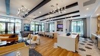 This single-floor amenity space presents a contemporary open-plan layout designed for both social gathering and relaxed lounging. The interior is unified by warm-toned hardwood flooring that flows throughout, complemented by crisp white walls and bold dark exposed ceiling beams that add architectural contrast. A series of striking, sculptural branch-style chandeliers are suspended overhead, providing ambient illumination alongside recessed lighting.

The space is organized into distinct functional zones. A generously sized lounge area features plush seating, including a mustard-toned sofa, upholstered armchairs, and glass-top accent tables, anchored by a textured stone-clad wall with a wall-mounted television. Adjacent to this is a long communal dining table surrounded by white modern chairs, ideal for gatherings or events.

A kitchen-style bar area with a white quartz countertop and light wood cabinetry serves the space, while a separate nook showcases hexagonal patterned floor tiles and geometric wood shelving, adding a decorative touch. Floor-to-ceiling black-framed windows and glass doors throughout flood the interior with natural light and offer lush tropical garden views, creating a seamless connection between the interior and the surrounding outdoor landscape.