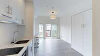 This modern single-floor space showcases a clean, open-concept design with a seamless flow between the kitchen and living areas. The interior is finished in a crisp, all-white palette complemented by light grey luxury vinyl plank flooring throughout, creating a bright and airy atmosphere. 

The kitchen is outfitted with sleek white cabinetry featuring stainless steel hardware, a light-toned stone countertop, and an undermount sink — offering both style and functionality. A small breakfast bar with seating provides a casual dining option that connects naturally to the living space.

The living area is bathed in natural light, courtesy of large sliding glass doors that open directly onto a private balcony, framing views of the surrounding cityscape. A stylish drum pendant light fixture anchors the living space, adding a warm focal point to the otherwise minimalist design.

The balcony, enclosed by a modern metal railing, extends the living space outdoors and offers an inviting spot to take in urban views. Throughout the unit, the combination of white walls, high ceilings, and abundant natural light creates a sense of openness and ease. The overall design is contemporary and understated, making it a highly functional and visually cohesive space.