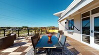 This single-story space combines functional interior design with an impressive outdoor terrace. The rooftop deck is a standout feature, finished with a distinctive checkerboard-patterned composite decking in alternating wood tones. It is outfitted with a glass-top dining table and seating alongside a separate lounge area furnished with yellow upholstered seating, offering expansive panoramic views of the surrounding landscape under open skies. Large glass doors connect the interior to the terrace, allowing abundant natural light to flow inward.

Inside, the space is defined by clean, modern finishes. Neutral gray walls are paired with wood-look luxury vinyl plank flooring throughout, creating a cohesive and contemporary aesthetic. A wide corridor with recessed ceiling lighting runs through the interior, lined with multiple gray panel doors fitted with brushed metal hardware. The hallway leads to a paneled entry door, reinforcing the structured layout of the space. Individual rooms feature smooth walls and consistent flooring, providing versatile, move-in-ready interiors. Overall, the space balances practical functionality with polished design details and notable outdoor living amenities.