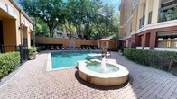 This inviting outdoor amenity space showcases a generously sized, curved swimming pool with crystal-clear turquoise water, designed to serve as the centerpiece of a well-maintained courtyard. A standout feature is the circular fountain spa positioned at the pool's edge, where a bubbling water jet creates a relaxing focal point and adds an element of resort-style sophistication. The pool deck is finished with classic brick paver hardscaping, providing a durable and visually appealing surface throughout the space. A row of lounge chairs with red cushions lines the perimeter, offering ample space for sunbathing and relaxation. Shaded seating areas with patio tables and vibrant red umbrellas are available for those seeking relief from the sun. A decorative arched stone wall feature serves as an elegant backdrop along one side. The surrounding mature trees provide natural shade and a lush, verdant canopy overhead. A charming auxiliary structure with a pitched tile roof, French-style windows, wall-mounted lanterns, and manicured shrubbery anchors one end of the courtyard, lending a refined, European-influenced character to the overall design. The multi-story building facade of warm golden stucco with balconies frames the space, creating an enclosed and private atmosphere ideal for leisure and outdoor enjoyment.