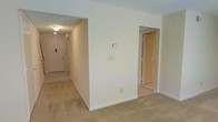 This two-story townhouse at Brookridge Village Apartments offers a straightforward, functional layout designed for comfortable everyday living. The main level features wall-to-wall carpeting in a neutral tone that carries throughout the living area, contributing to a warm and understated atmosphere. Crisp white walls and white baseboards keep the space feeling clean and bright, while a flush-mounted ceiling light provides consistent overhead illumination.

A defining feature of the main level is the partial wall that divides the interior, creating a natural separation between the entryway corridor and the adjoining living space. The front entry area is accessible directly from the main living zone, with a solid exterior door fitted with a deadbolt for added security. An additional doorway on the opposite side of the partial wall leads to a secondary room or hallway, enhancing the overall flow and connectivity between spaces.

Electrical outlets and switch plates are thoughtfully positioned along the walls, supporting practical furnishing arrangements. The two-story configuration provides additional living or sleeping space on the upper level, making this townhouse a versatile option within the Brookridge Village community.