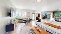 Located at 5855 Old Oak Ridge Rd in Greensboro, NC, this thoughtfully designed single-floor community space offers a generous, open layout that seamlessly connects multiple functional areas. The interior is finished with light wood-look flooring throughout, creating a warm and cohesive aesthetic across all zones.

The living room is a bright, welcoming area anchored by a large sofa, mid-century modern coffee table, and an accent wall painted in a soft sage green. Natural light floods the space through wide windows, offering views of the surrounding greenery. A wall-mounted TV and stylish cabinetry round out the room's functionality.

The combined dining and lounge area is a design highlight, featuring a bold teal subway tile accent wall with open shelving above dark cabinetry and granite countertops. A striking dining table with a wood-inlay top is illuminated by a cluster of globe pendant lights, creating an inviting atmosphere for gathering.

A dedicated lounge zone showcases a deep navy blue feature wall, curved banquette seating, and circular tables ideal for casual meetings or relaxed conversation. Adjacent to this area, built-in workstations with leather seating and monitor setups provide practical workspace. Curated artwork, greenery, and layered geometric rugs tie the entire space together with a polished, contemporary feel.